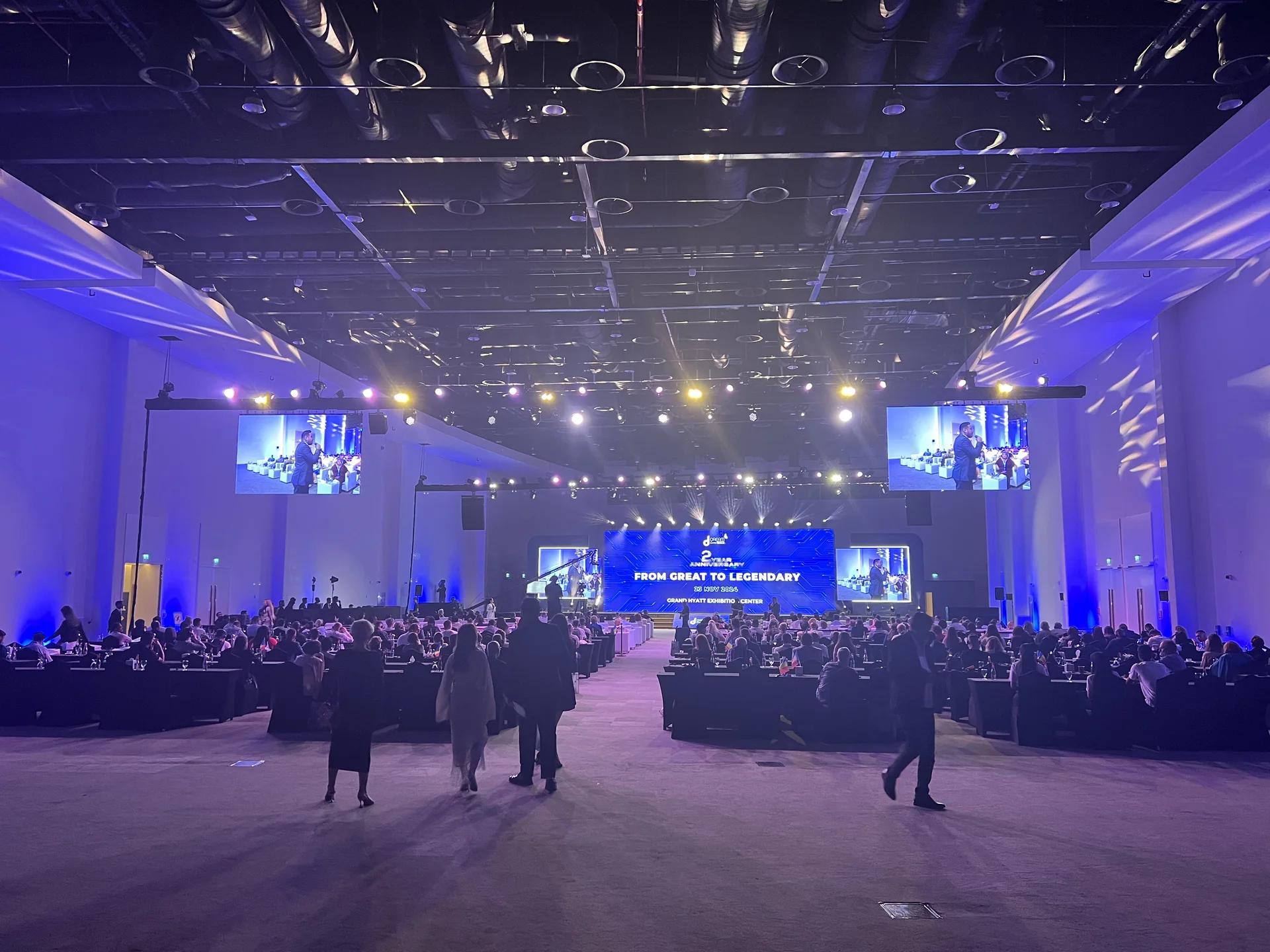 Large conference hall with delegate seating, an illuminated stage and suspended screens.
