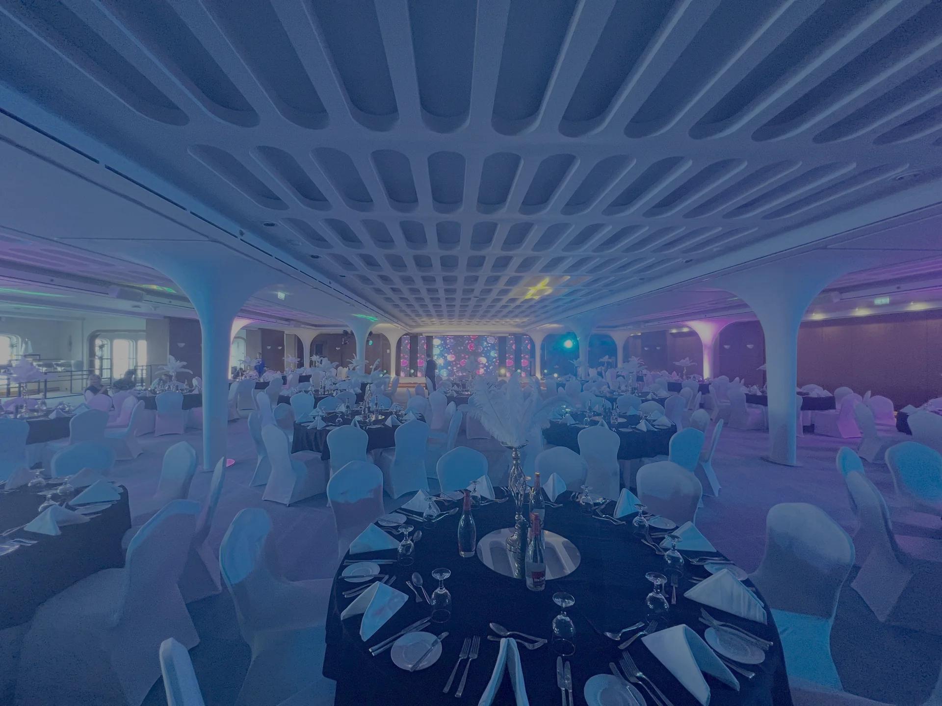 Banquet room with guest tables, stage lighting and a distant presentation area.