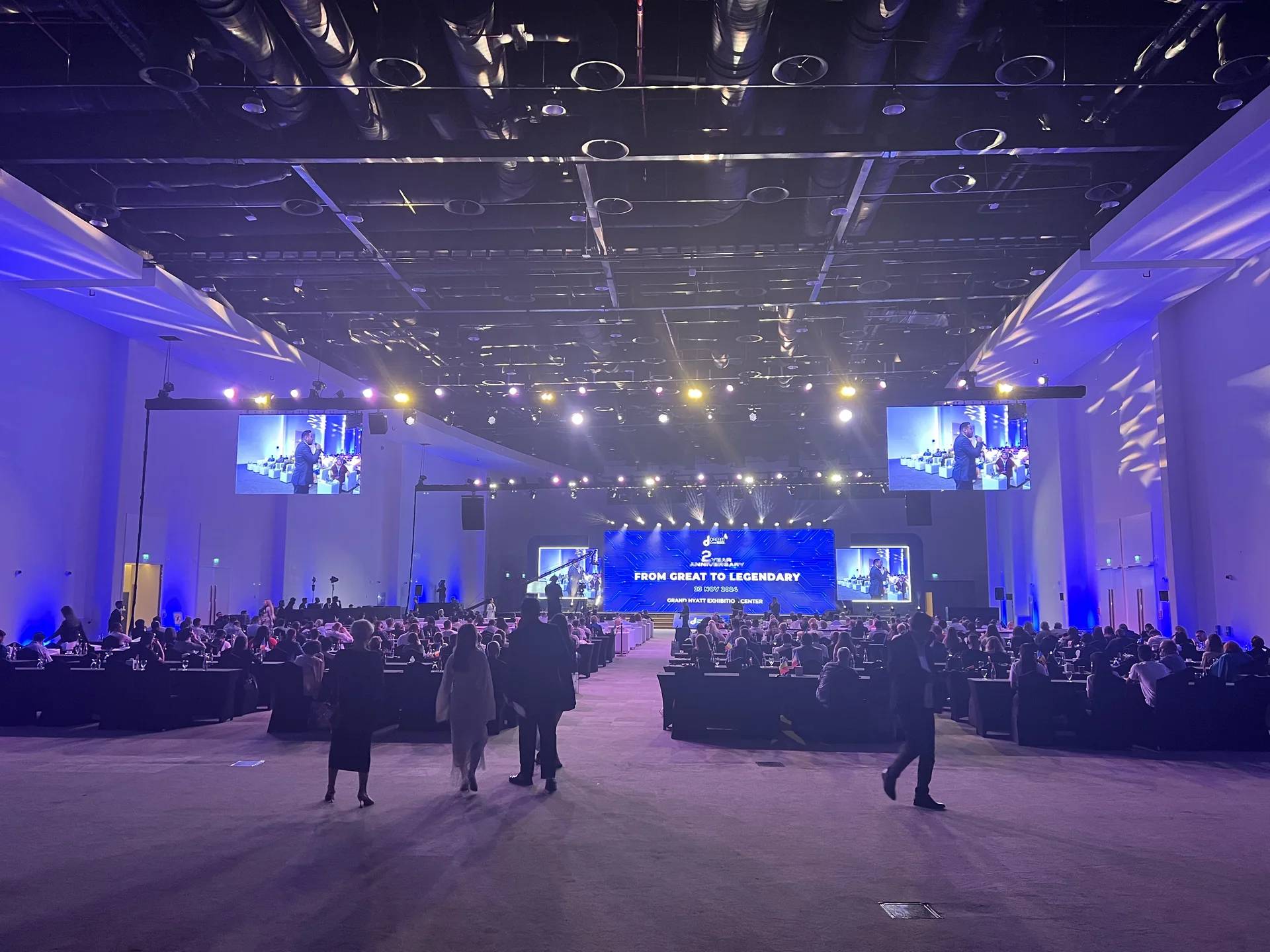 Large conference hall with delegate seating, an illuminated stage and suspended screens.